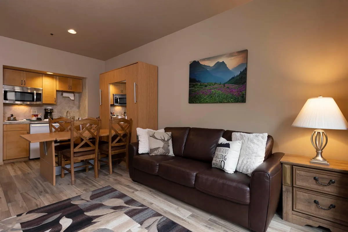 A warm and inviting living space with a leather sofa, a dining table set for two, and a large mountain landscape artwork on the wall. Soft lighting from a table lamp enhances the cozy atmosphere, perfect for relaxing or sharing intimate moments together.