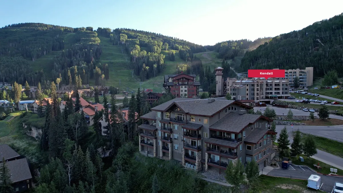 A wide view of the resort's exterior with mountain slopes in the background, hotel buildings nestled among trees, and a visible parking area. The scene conveys both adventure and tranquility, showing the proximity to nature and outdoor activities.