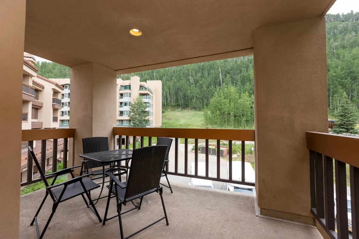 This exterior image shows the inviting outdoor seating area surrounded by lush greenery, with the building architecture visible in the background. The setting conveys relaxation and social connection, suggesting easy access to communal village spaces, perfect for enjoying morning coffee, meeting friends, or unwinding after a day of adventure.