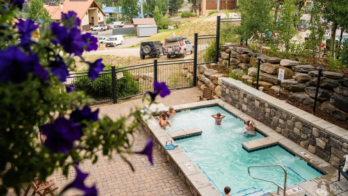 A lively outdoor scene featuring a hot tub filled with guests laughing and relaxing, surrounded by colorful flowers and resort landscaping. Children splash in the pool nearby, illustrating the fun and social aspect of the shared amenities.