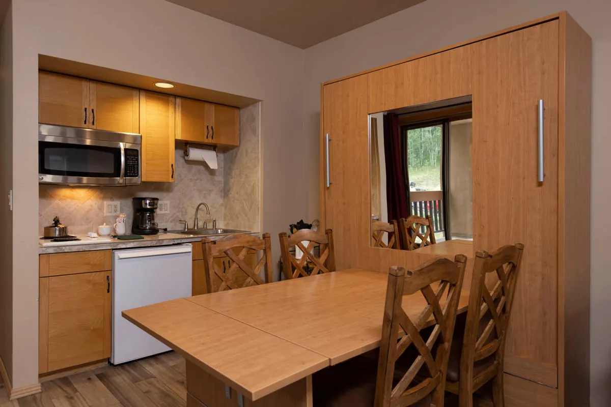 A view into the full kitchen and adjacent dining area, highlighting kitchen appliances, cabinets, and a spacious table. The setup encourages preparing meals together and enjoying them in the heart of the home.