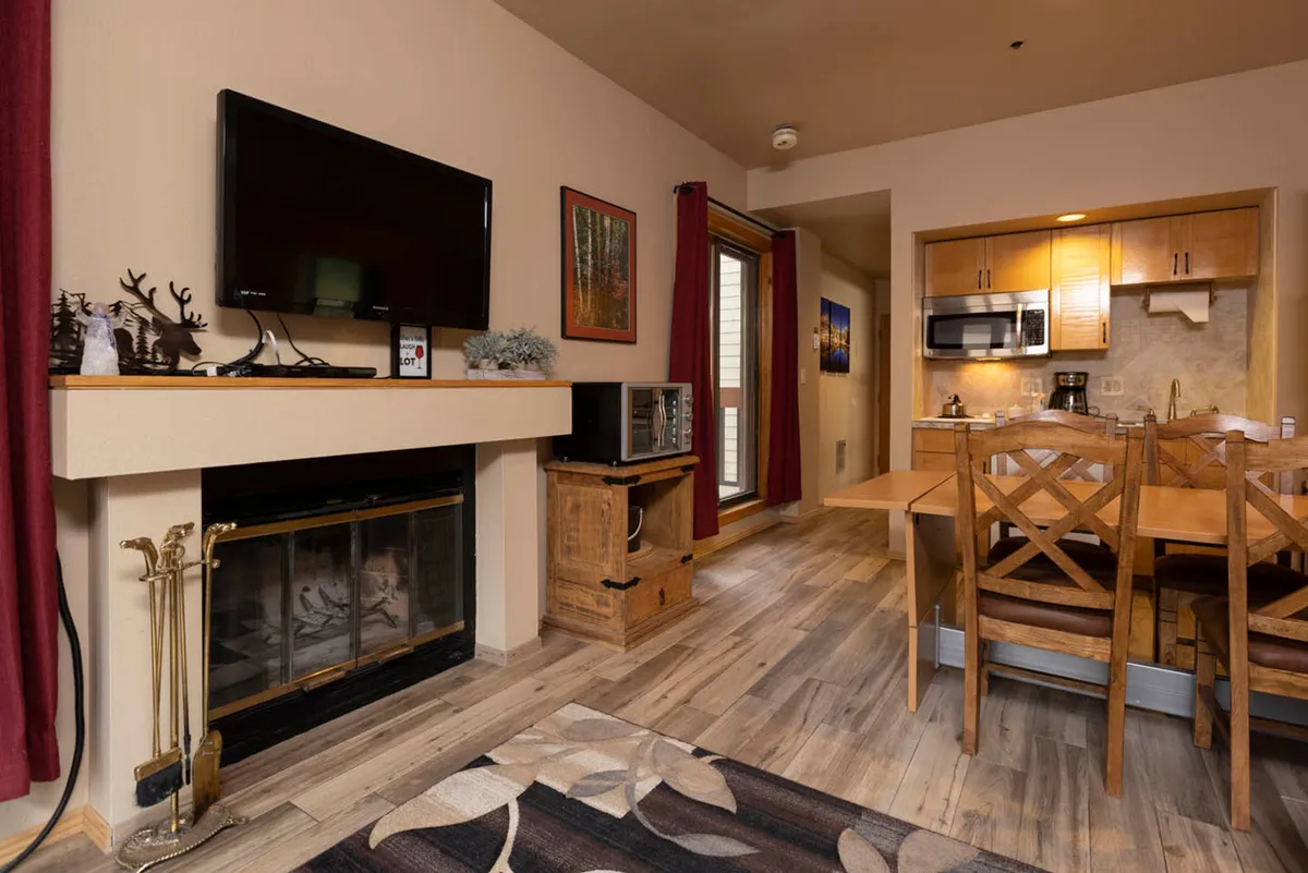 The image showcases a cozy condo interior with a crackling wood-burning fireplace as the centerpiece, a welcoming dining table set for meals, and a fully equipped kitchen visible in the background. The space is designed for relaxation and connection, perfect for unwinding after mountain adventures. Soft lighting and comfortable furnishings create a warm, inviting ambiance.