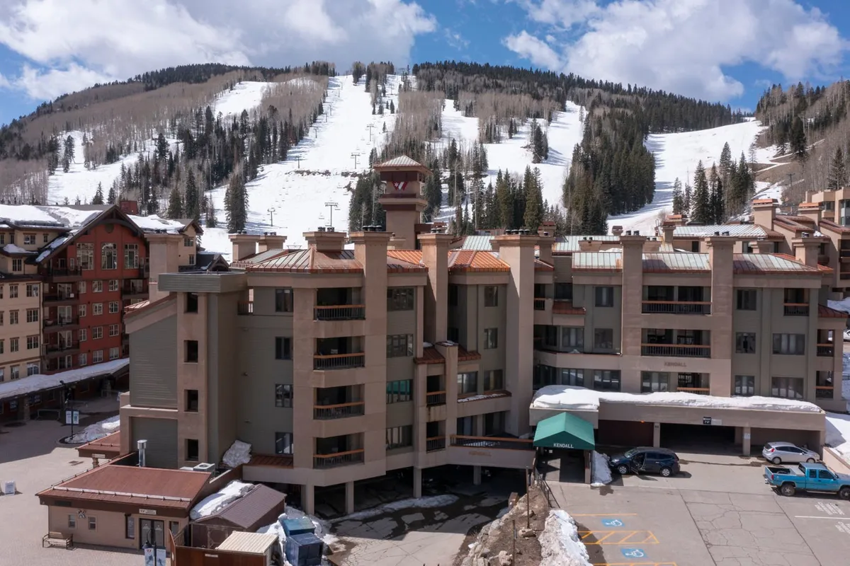 An energetic exterior shot of the property's multilevel building with the surrounding ski slopes and parking area visible. The scene is lively, with the slopes beckoning in the background, and the parking area confirming the availability of convenient garage access for guests.