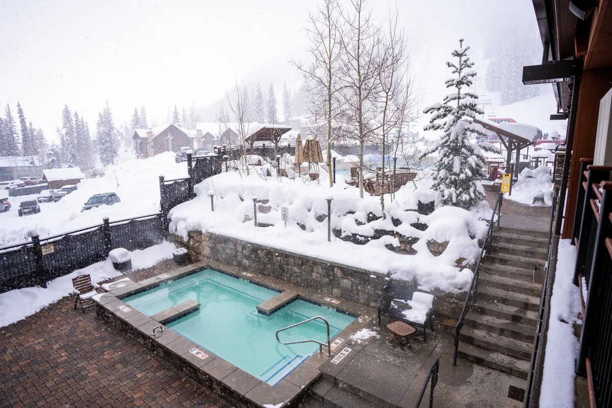 An inviting heated outdoor pool surrounded by snow-covered trees and a mountain backdrop. Comfortable seating areas line the pool, and a hot tub steams gently in the cool air. The entire setting exudes tranquil luxury and captures the unique appeal of swimming and soaking outdoors in any season, especially in winter.