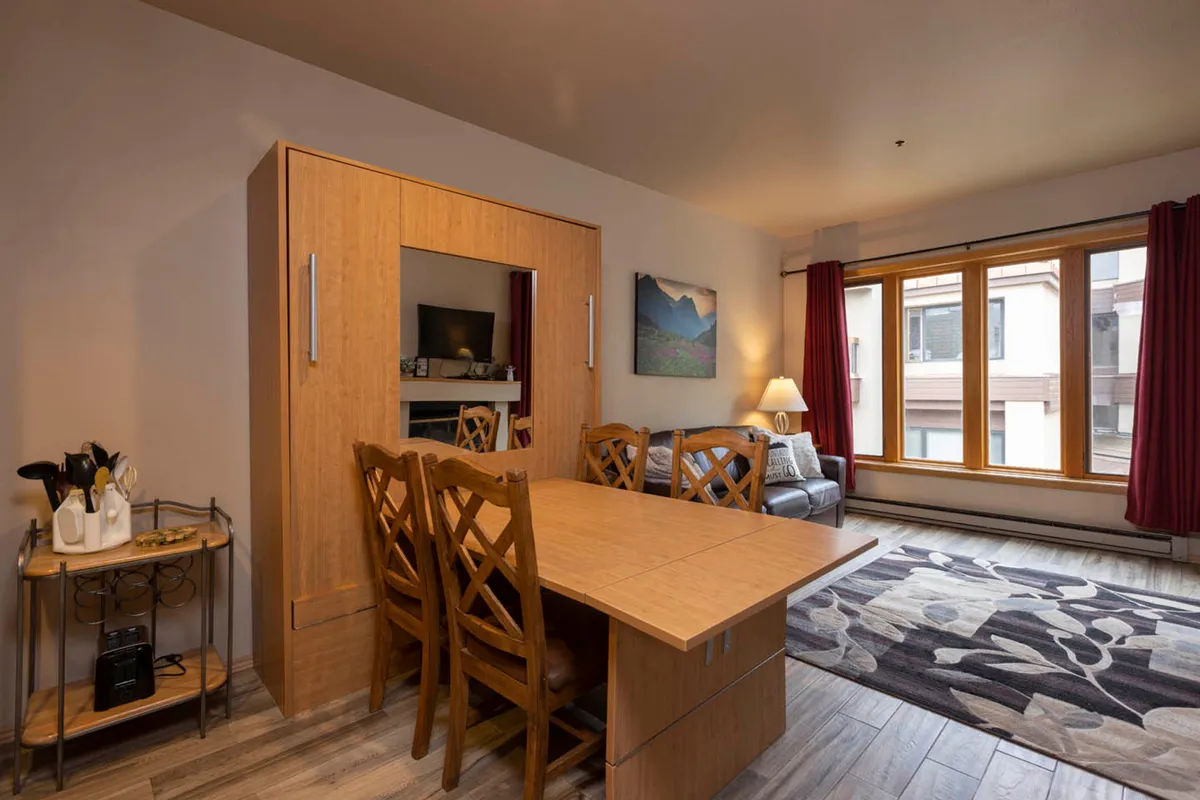 A warm, well-lit interior revealing an open-plan setup with a dining table, comfortable sofa, large windows, and a decorative rug. The space is ideal for family meals, casual gatherings, or simply relaxing while enjoying the view.