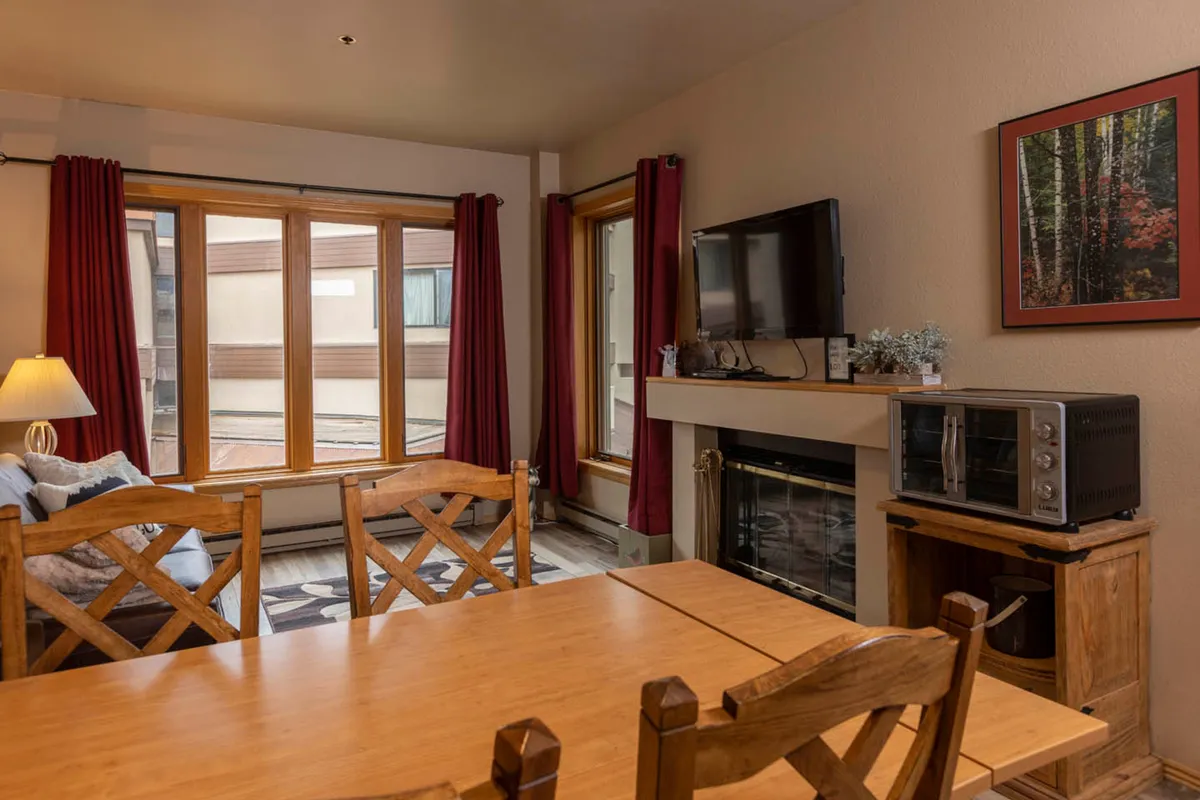 A spacious, inviting living and dining area featuring a prominent wood-burning fireplace, large windows that fill the room with natural light, a cozy dining table, and a television. The space radiates warmth and encourages relaxation, gathering, and connection.