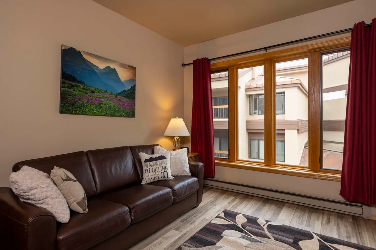 A cozy interior view featuring a leather sofa, a large window with red curtains, a table lamp, and mountain landscape artwork. The space feels warm and welcoming, ideal for relaxing, socializing, or remote work, with soft natural light hinting at the vibrant world just outside.