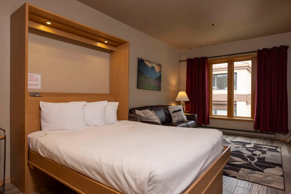 A cozy interior bedroom scene featuring a plush bed with soft linens, a window letting in natural light, a comfortable sofa, and a warm lamp. The arrangement suggests a relaxing space ideal for winding down, with thoughtful decorative touches.